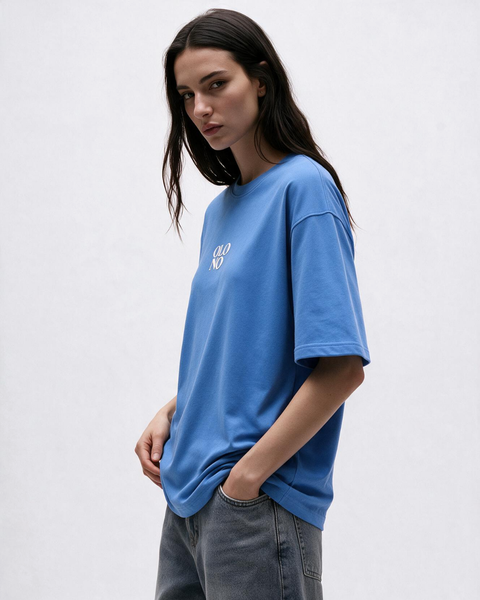 A woman wearing a blue olono oversized short-sleeve t-shirt with a small white graphic on the left chest, paired with gray jeans, against a plain light gray background.