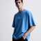 A man wearing a light blue oversized t-shirt with a small white 'OLONO' logo on the chest, paired with black pants, photographed against a light gray studio background.