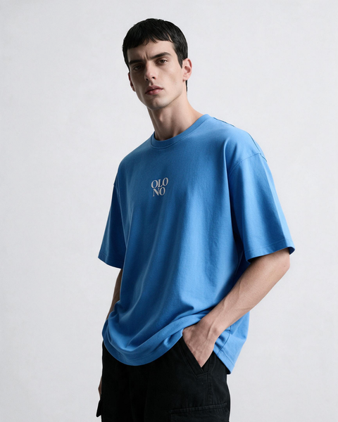A man wearing a light blue oversized t-shirt with a small white 'OLONO' logo on the chest, paired with black pants, photographed against a light gray studio background.