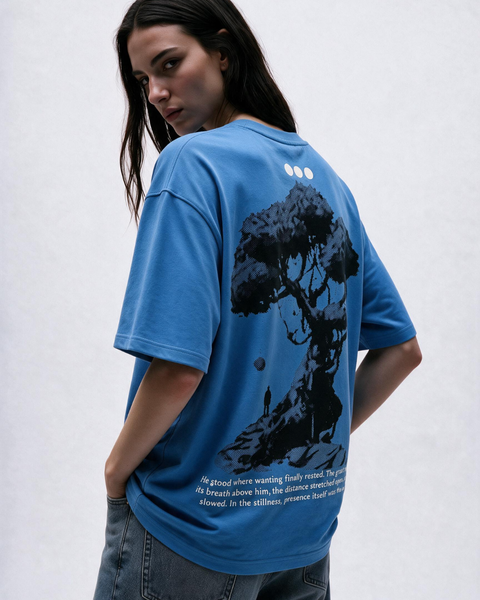 A woman wearing an Olono oversized blue graphic T-shirt, shown from the back. The shirt features a large black tree illustration and white text on the back, paired with gray jeans against a light gray background.