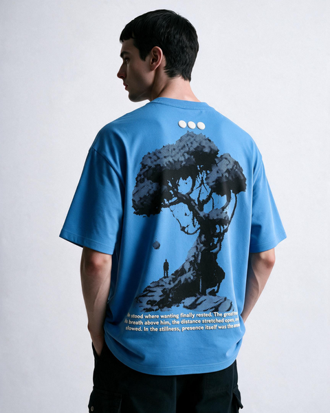 A man wearing a light blue Olono oversized T-shirt, shown from the back. The shirt features a large black graphic of a tree on a hill with a small figure, three white circular details at the neck, and white text at the bottom.