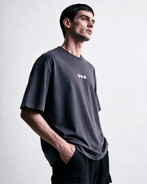 A man wearing a dark charcoal gray oversized t-shirt with three small light-colored dots on the left chest, paired with black pants, photographed against a neutral light gray background.