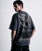 A man wearing a dark charcoal gray oversized t-shirt with a large white 'SILENCE' graphic and a black-and-white photo print on the back, shown from a three-quarters back view against a light gray studio background.
