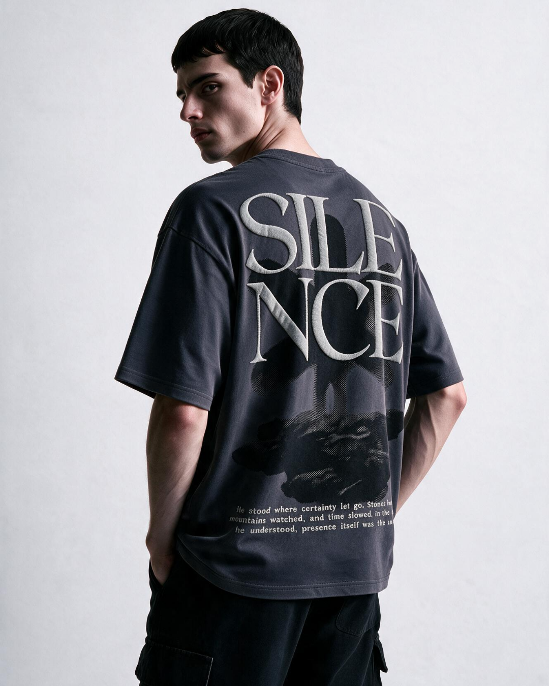 A man wearing a dark charcoal gray oversized t-shirt with a large white 'SILENCE' graphic and a black-and-white photo print on the back, shown from a three-quarters back view against a light gray studio background.