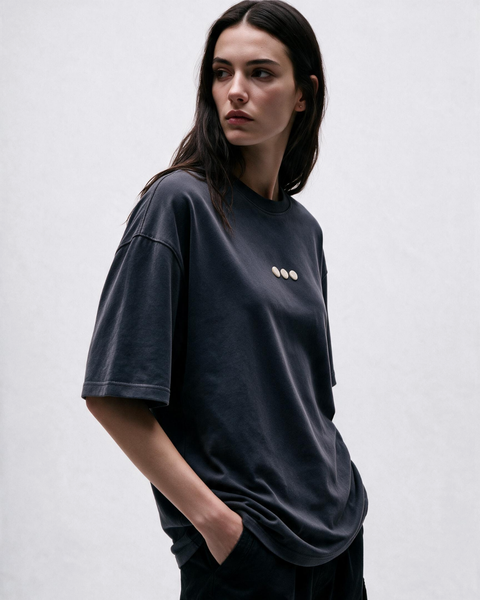 A woman wearing a dark charcoal gray oversized t-shirt with three small gold buttons on the left chest, posed against a light gray studio background.