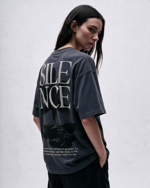 A woman wearing a dark charcoal gray oversized graphic T-shirt, shown from the back. The shirt features the word 'SILENCE' in large, stylized white letters with a black distressed graphic underneath and small white text near the hem.